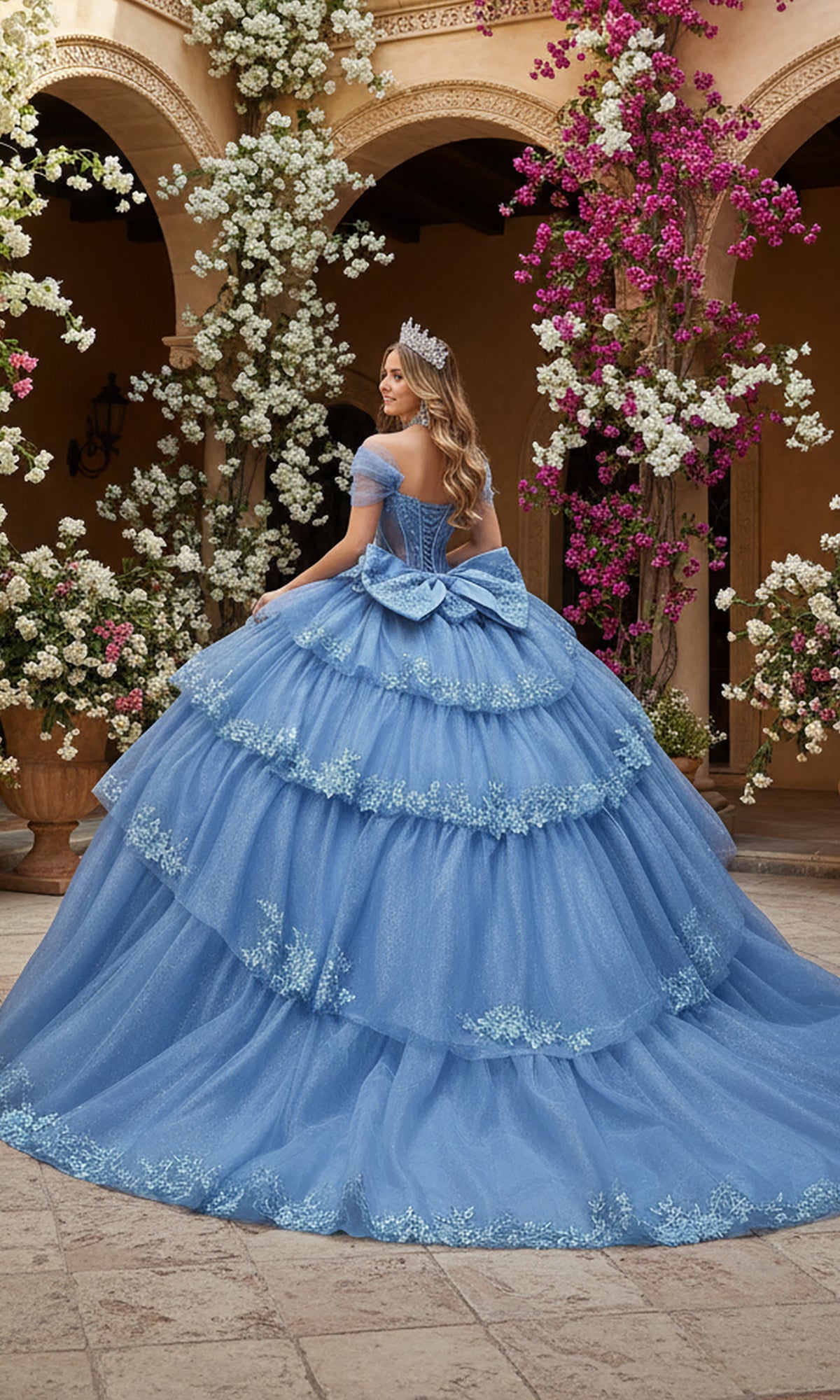 A woman in the Quinceanera Dress 5156 By Dancing Queen, featuring a blue lace gown with a sheer corset bodice, stands in a courtyard of white and pink flowers, her sparkling tiara completing the elegant look.